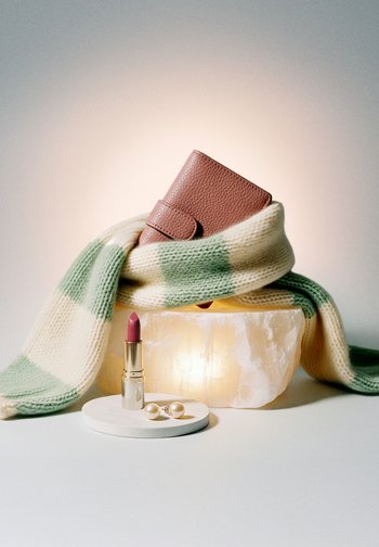 Brązowy portfel o fakturze, owinięty zielono-kremowym dzianinowym szalikiem, różowa szminka i perłowe kolczyki na białym tle z miękkim oświetleniem.