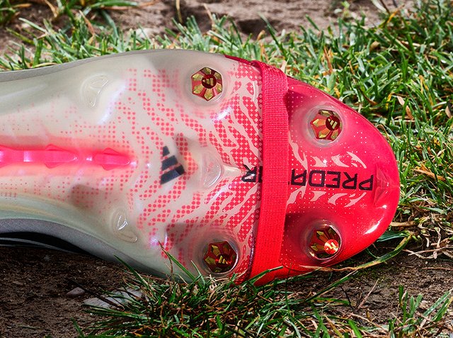 Piros-fehér Adidas Predator focicipő tiszta talpa fém stoplikkal, fű és föld felületen.