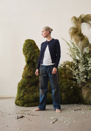 Een persoon staat blootsvoets in een mosachtige omgeving, gekleed in een marineblauwe cardigan en versleten jeans, omringd door bloemenarrangementen en puin.