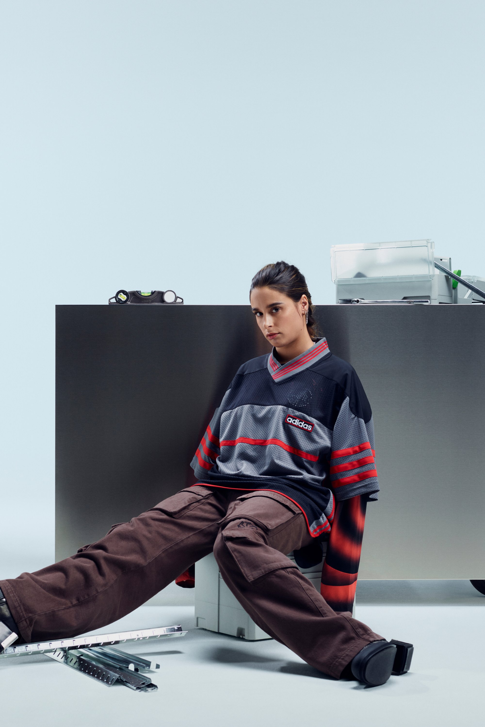 Een persoon zit casual tegen een metalen oppervlak, gekleed in een oversized shirt met patroon en cargobroeken, omringd door gereedschap en voorwerpen.