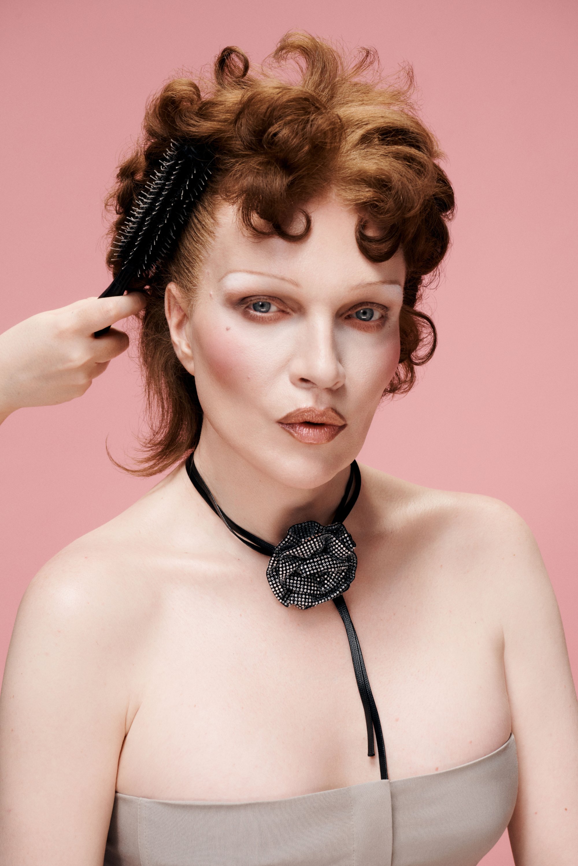 Una persona con cabello rizado y rojo y un maquillaje atrevido mira a la cámara, mientras una mano peina su cabello con un peine negro contra un fondo rosa.