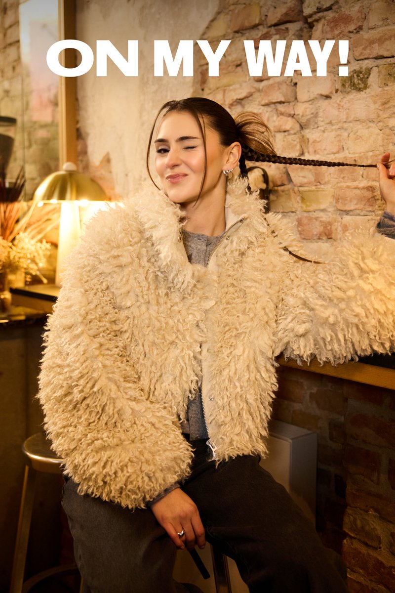 Ung kvinde i en fluffy frakke blinker og leger med sit fletninger hår foran en rustik murstensvæg med teksten "ON MY WAY!" ovenover.