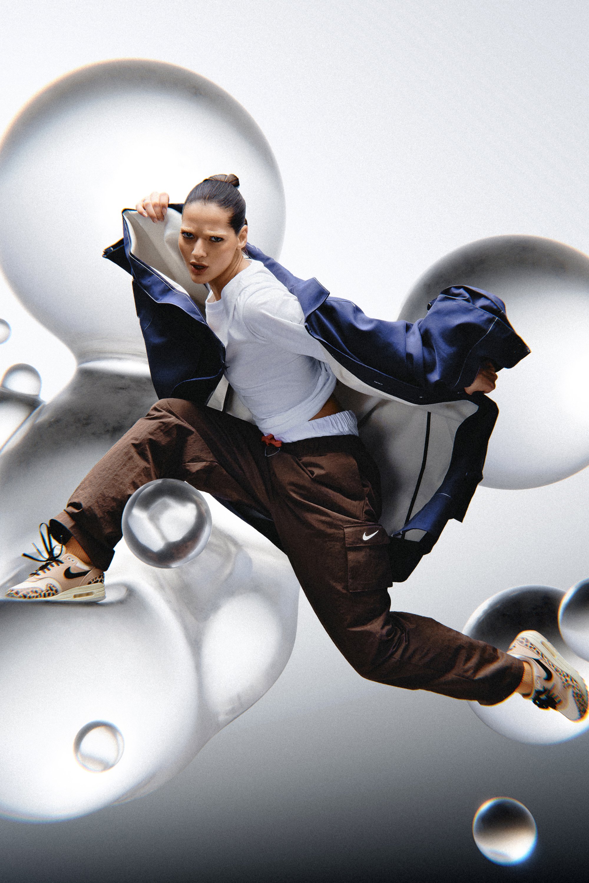 Een vrouw in een witte top en een bruine broek springt energiek, gekleed in Nike-schoenen, met abstracte bubbels in een dynamische, minimalistische achtergrond.