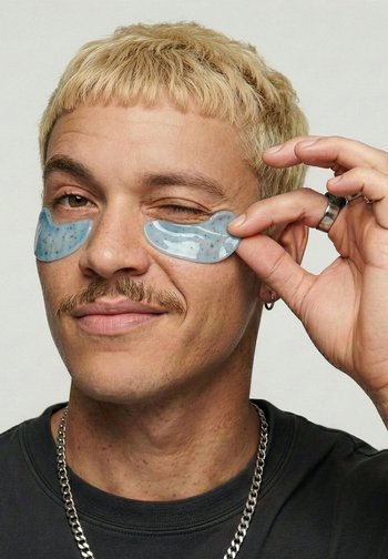 Lyshåret mand med overskæg påfører blå gel-patches under øjnene, vinker med det ene øje, iført en halskæde og mørk skjorte.