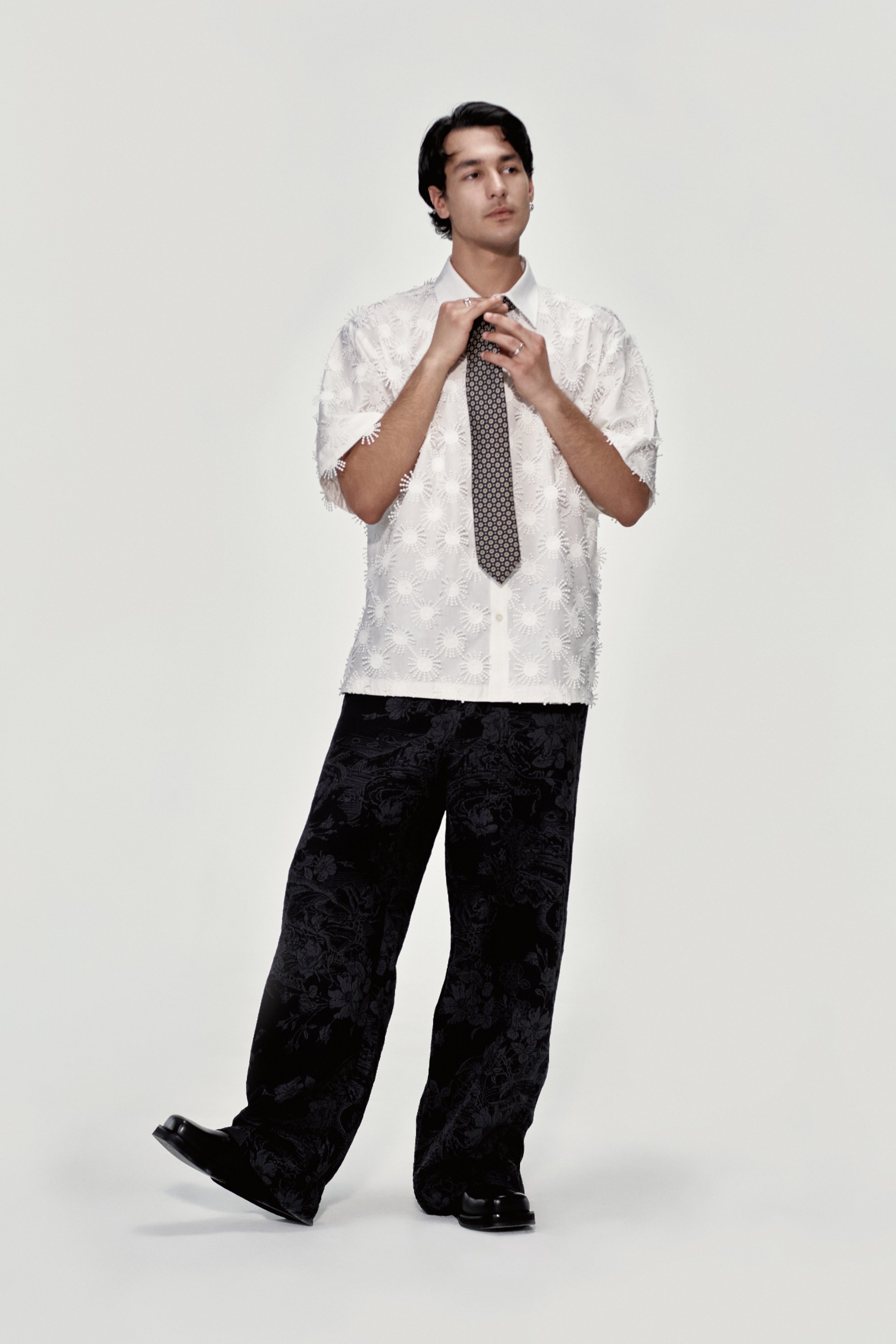 Un giovane in una camicia bianca decorativa e una cravatta a fantasia sta in modo pensieroso, indossando pantaloni neri larghi e scarpe, su uno sfondo semplice.