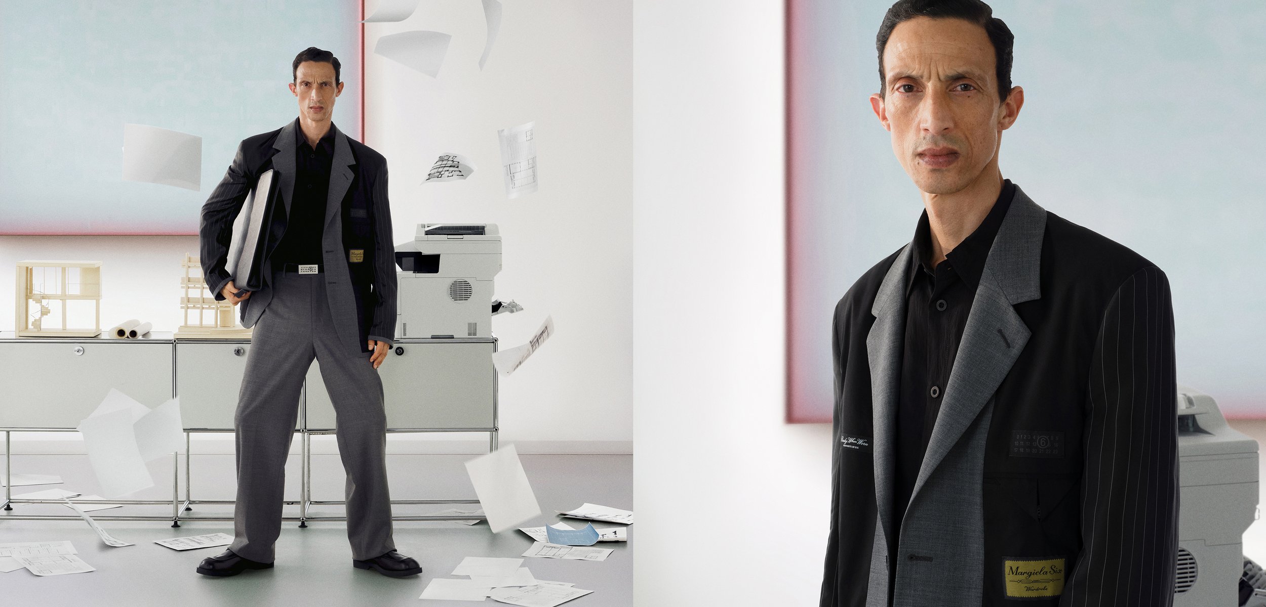 Vīrietis tumšā uzvalkā stāv pārliecināti birojā ar izkaisītām papīra lapām pie printera un rakstāmgalda, ar mierīgu izteiksmi, mūsdienīgā minimālisma stilā.