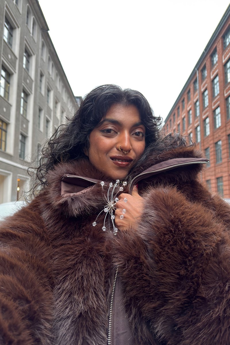 Person with curly black hair and nose rings wearing a brown fur coat with a crystal brooch, standing between tall gray and red brick buildings.