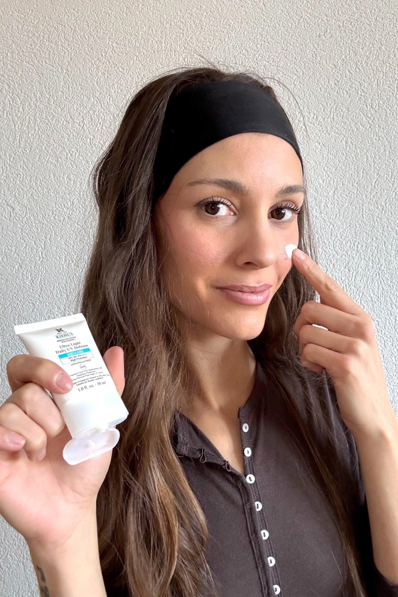 Donna con una fascia nera sulla testa che applica la crema solare Kiehl’s Ultra Light Daily UV Defense sulla guancia, con uno sfondo di parete semplice.