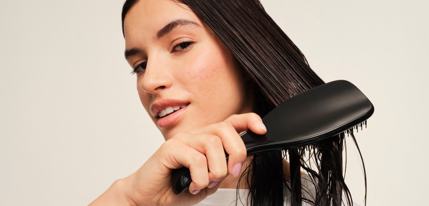Jeune femme brossant ses longs cheveux noirs mouillés avec une grande brosse plate noire sur un fond uni.