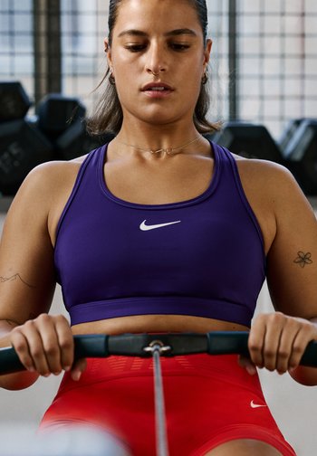 Violetti urheilullinen crop-top valkoisella Nike-logolla, yhdistettynä punaisiin korkeaotsaisiin shortseihin. Lähikuva käsistä, jotka pitävät kiinni soutulaitteen kahvasta.