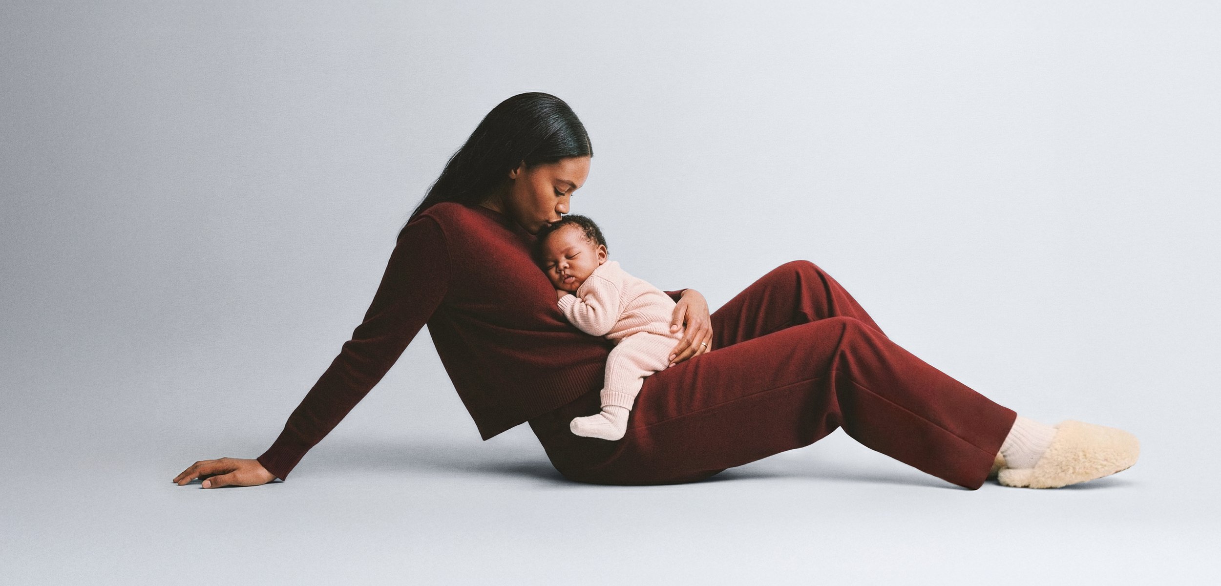Jersey y pantalones burdeos llevados por un adulto, sosteniendo un bebé con un conjunto rosa claro, sobre un fondo gris liso.