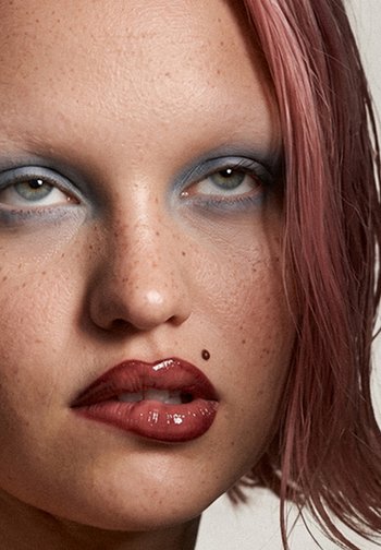 Gros plan d'une personne avec des cheveux roses, un fard à paupières bleu, un rouge à lèvres rouge foncé brillant et un petit grain de beauté au-dessus de la lèvre supérieure.