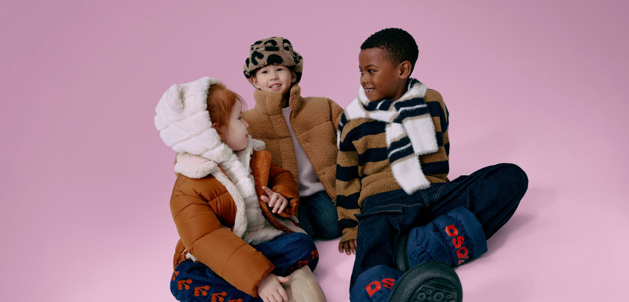 Τρία παιδιά καθισμένα μπροστά σε ένα ροζ φόντο. Φορούν πολύχρωμα χειμερινά ρούχα: ένα μπεζ φουσκωτό μπουφάν, ένα κρεμ παλτό και ένα ριγέ πουλόβερ.
