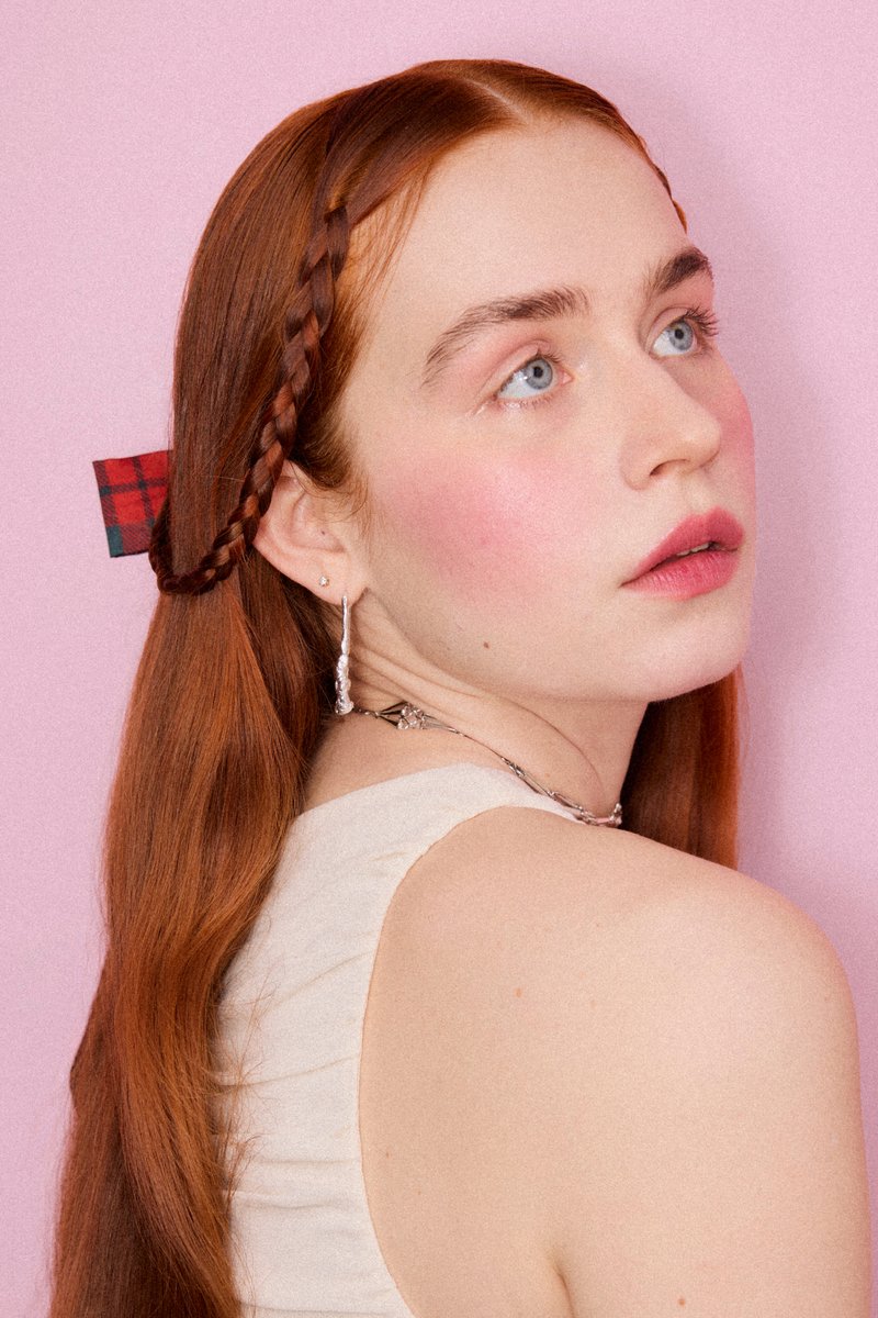 Rothaarige Frau mit geflochtenem Haar und karierter Schleife, trägt silberne Ohrringe und Halskette, schaut über ihre Schulter vor einem rosa Hintergrund.