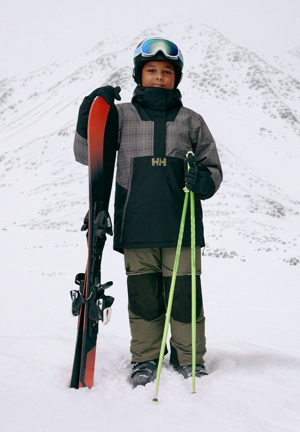 Snowboard med et rødt og svart design. Modellen har på seg en svart og gråmønstret jakke, grønne snøbukser og holder grønne skistaver i snøen.