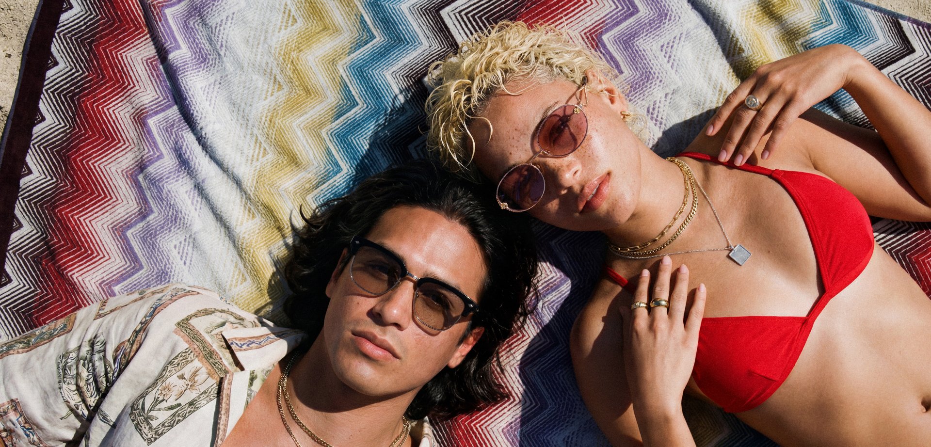 Δύο άτομα με γυαλιά ηλίου και κοσμήματα ξαπλώνουν σε μια πολύχρωμη κουβέρτα με ζιγκ-ζαγκ, το ένα φορώντας κόκκινο πάνω μέρος μπικίνι και το άλλο ένα μπουφάν με σχέδιο.