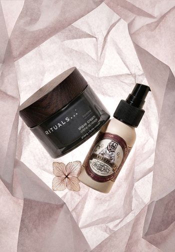 Frasco de crema de afeitar de Rituals y botella de aceite para barba de Mr. Bear Family colocados sobre papel translúcido arrugado con una flor seca.