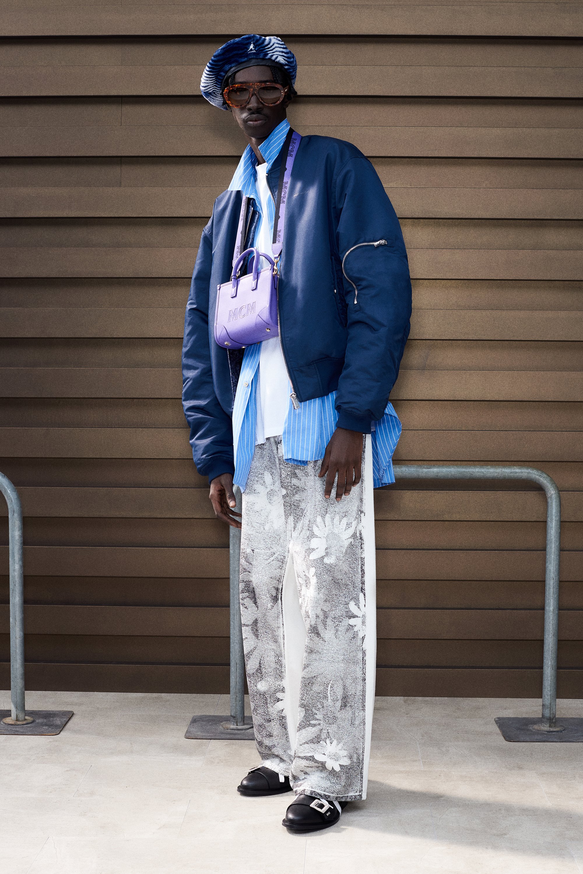 A tall model poses against a brown wall, wearing a navy bomber jacket, striped shirt, floral pants, and a purple handbag, with sunglasses and a hat.