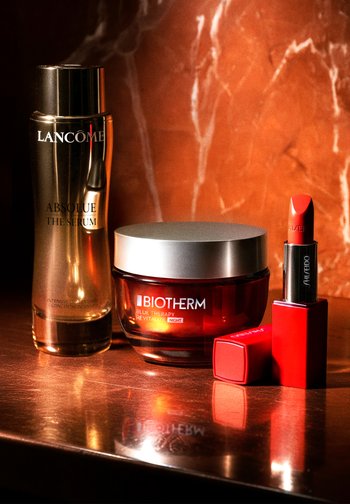Lancome gold serum bottle, Biotherm red cream jar with silver lid, and two Shiseido red lip products on a reflective surface against a marble backdrop.
