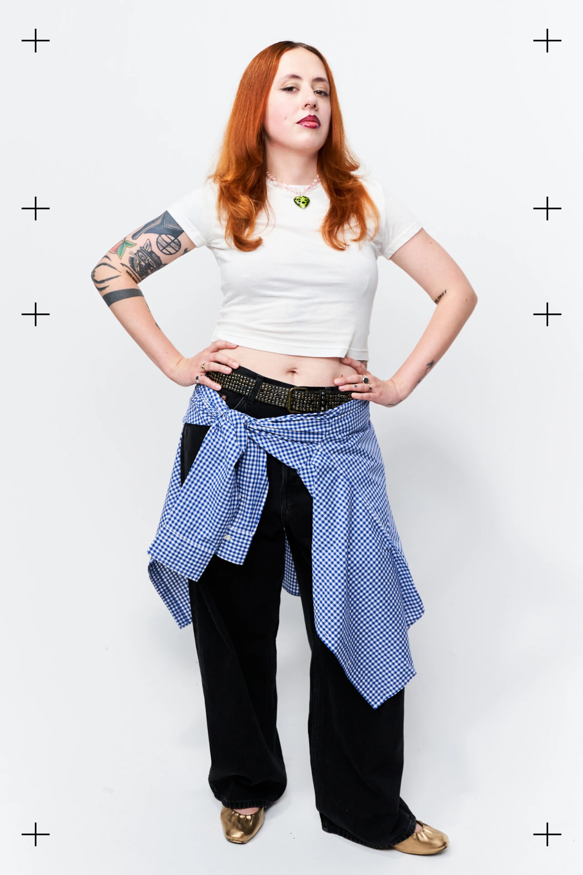 Una mujer con cabello rojo largo posa con confianza en una camiseta blanca corta, pantalones anchos negros y una camisa de cuadros azul atada a la cintura.