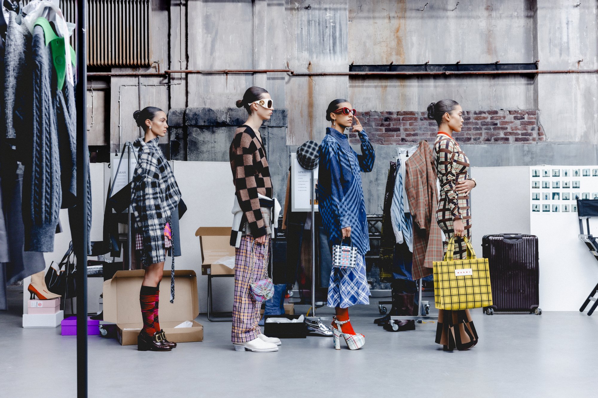 Five models showcase diverse fashion styles in a studio, surrounded by clothing racks and boxes, with a stylish backdrop.