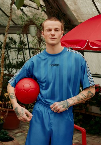 Junger Mann in blauer Adidas-Sportbekleidung zwinkert, hält einen roten Fußball und steht in einem Gewächshaus mit Pflanzen und einem roten Regenschirm im Hintergrund.