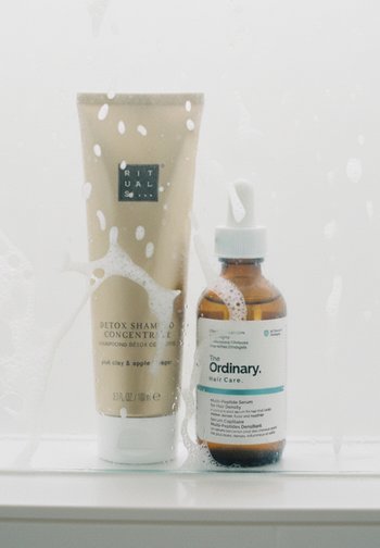 Champú detox ritual en un tubo mate de crema y suero The Ordinary en una botella de vidrio oscura, ambos en un entorno de ducha con espuma.