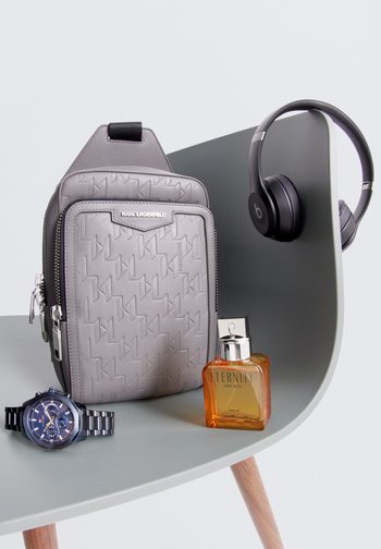 Graue Karl Lagerfeld Tasche, schwarze Kopfhörer, blaue Uhr und Calvin Klein Eternity Duft auf einem geschwungenen grünen Stuhl.