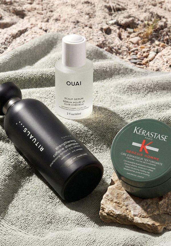 Schwarze Flasche mit Koffeinshampoo, weiße Flasche mit Kopfhauttserum und grüner Haarverdickungs-Tonbehälter auf strukturiertem grünem Handtuch im Freien.
