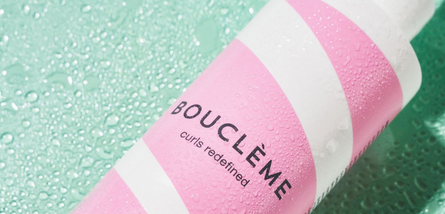 Nahaufnahme einer rosa-weiß gestreiften Bouclème-Haarpflegeflasche mit Wassertropfen auf einer grünen Oberfläche.
