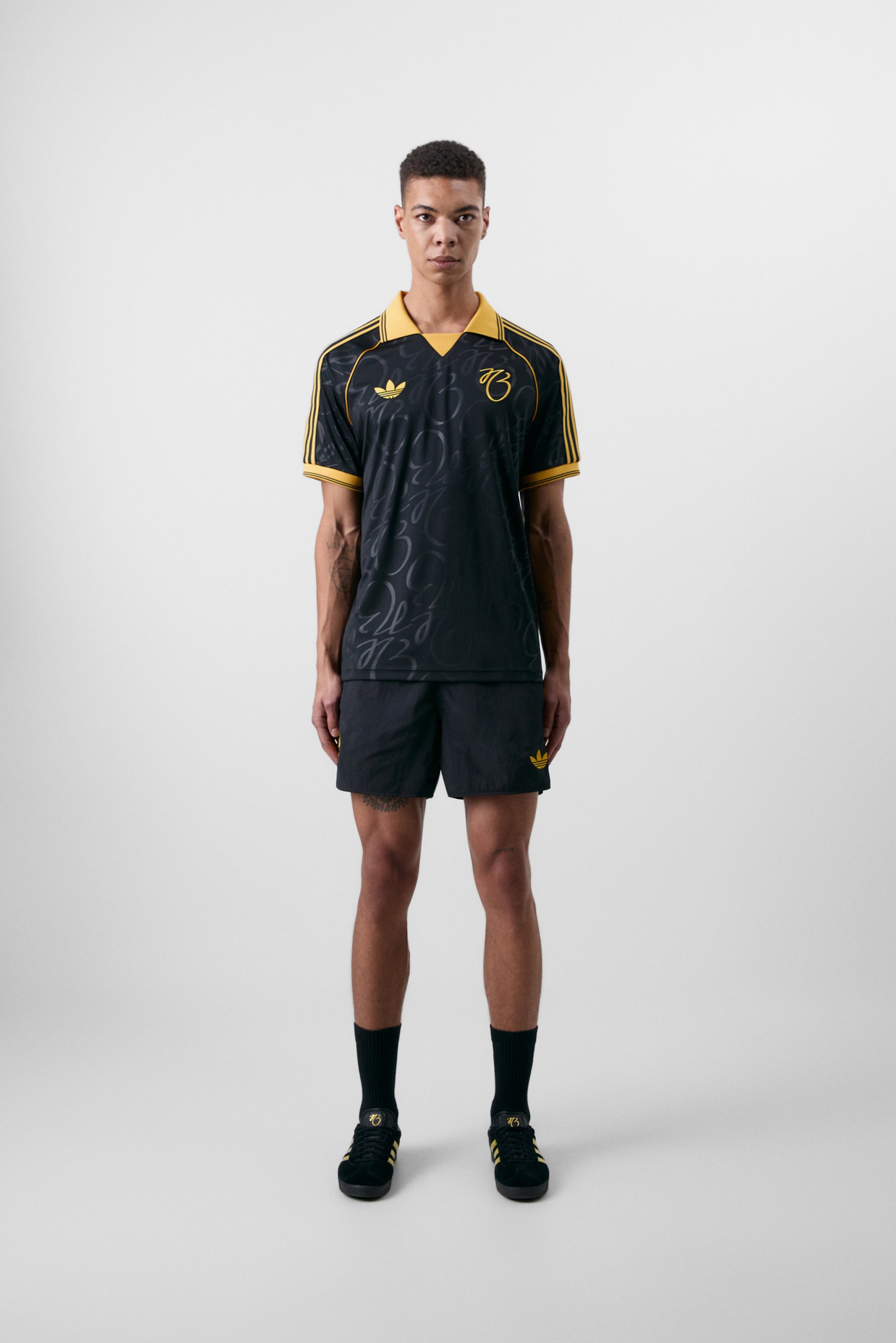 Un joven modelo masculino está de pie, vistiendo una camiseta deportiva negra y amarilla con pantalones cortos y zapatillas a juego, frente a un fondo neutro.