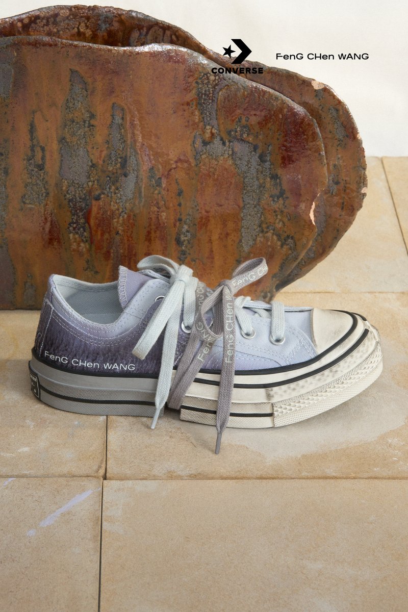 Grey and white Converse trainers with distinctive laces rest on a textured surface, set against a backdrop of rust-coloured decorative elements.