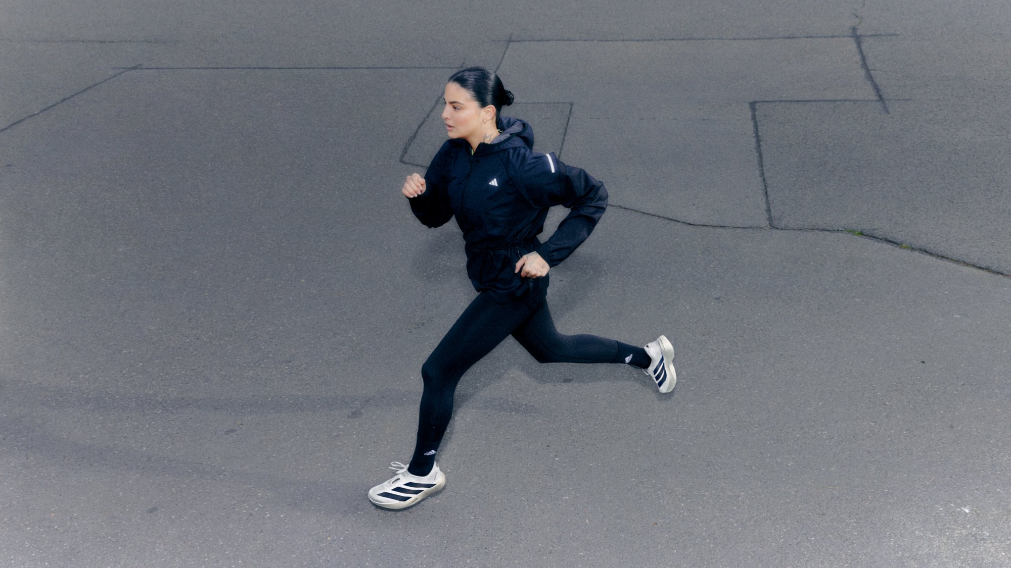 Una persona con una chaqueta deportiva negra y leggings corre sobre una superficie de asfalto gris, mostrando una pose activa y atlética.