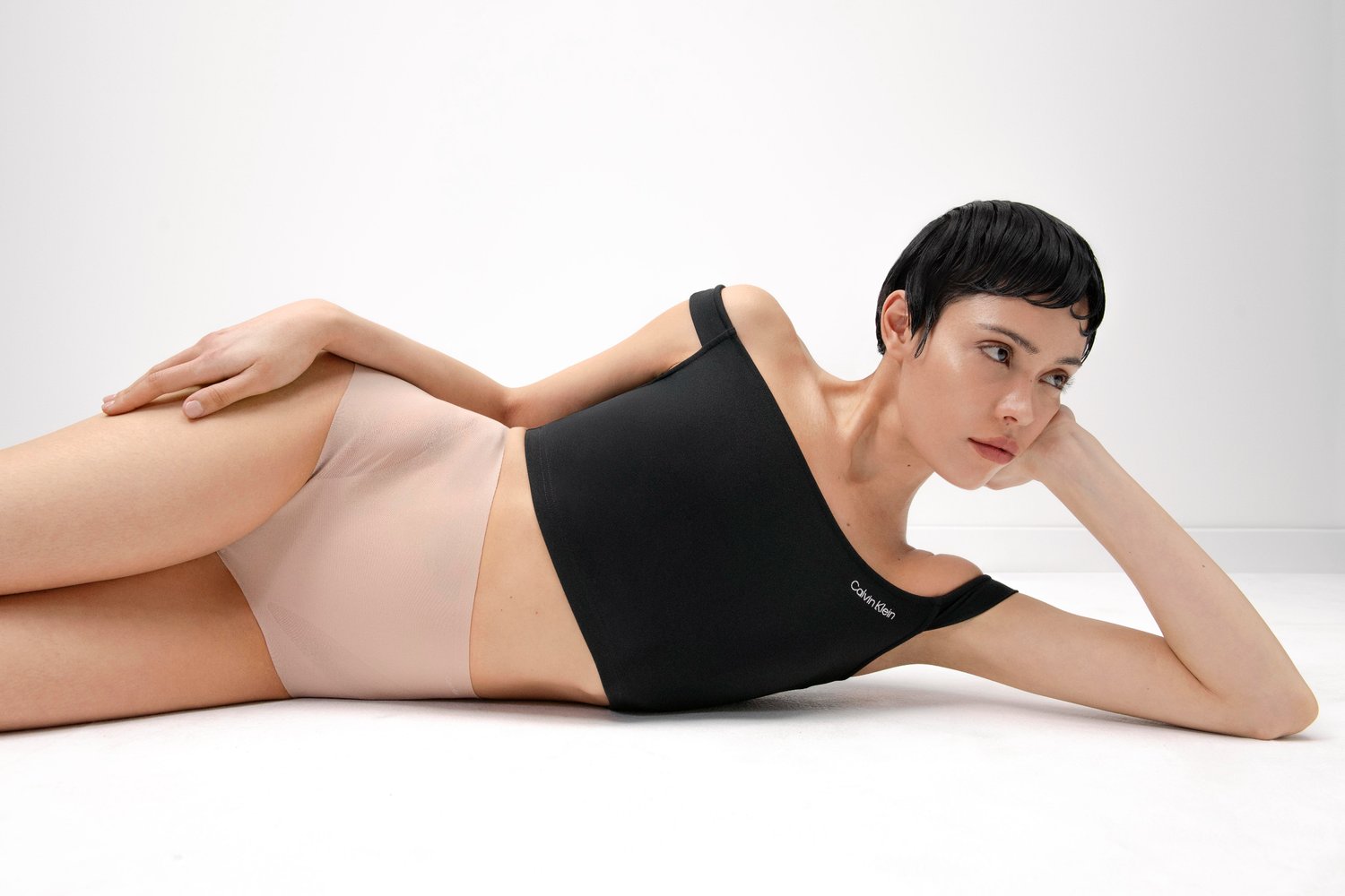 Model ligt ontspannen op de witte vloer, gekleed in een zwarte crop top en huidskleurige ondergoed, met haar kin op haar hand gesteund en een contemplatieve uitdrukking.