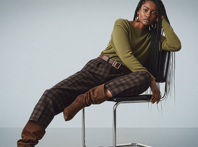 Grön stickad tröja, rutiga bruna byxor med hög midja, bruna knähöga stövlar och ett brunt bälte. Modell sitter på en metal stol med flätor.