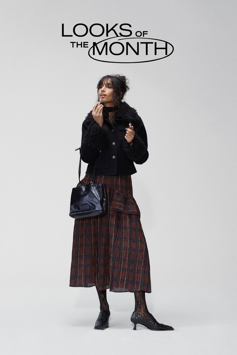 Zwarte cropped jas met een pluizige kraag, geruite rok in diep rood en bruin, kanten panty’s en zwarte puntige hakken, terwijl ze lipstick vasthoudt.