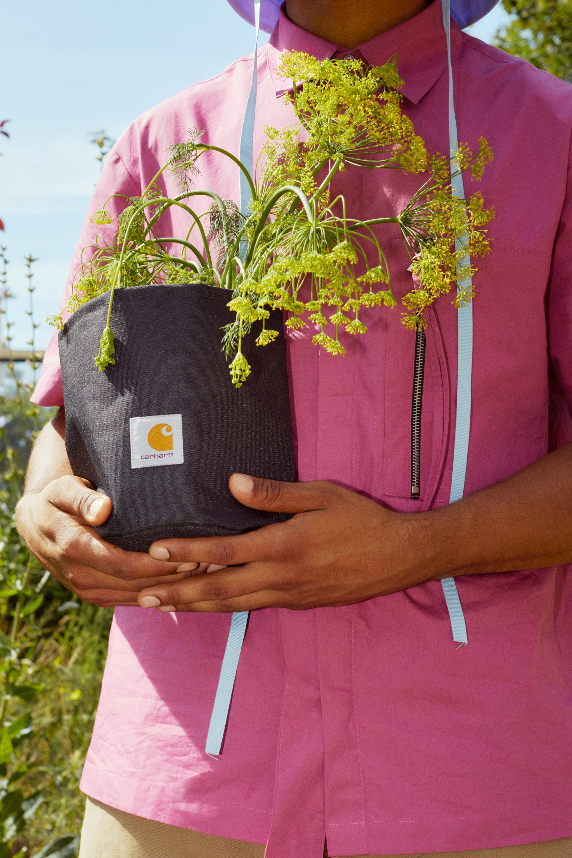 Una persona vestida con una camisa rosa sostiene una maceta negra con plantas verdes y florecientes, frente a un fondo de vegetación y cielo azul.