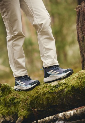 Person som har på seg beige bukser og svarte tursko, går på en mosegrodd stok i skogsområde.