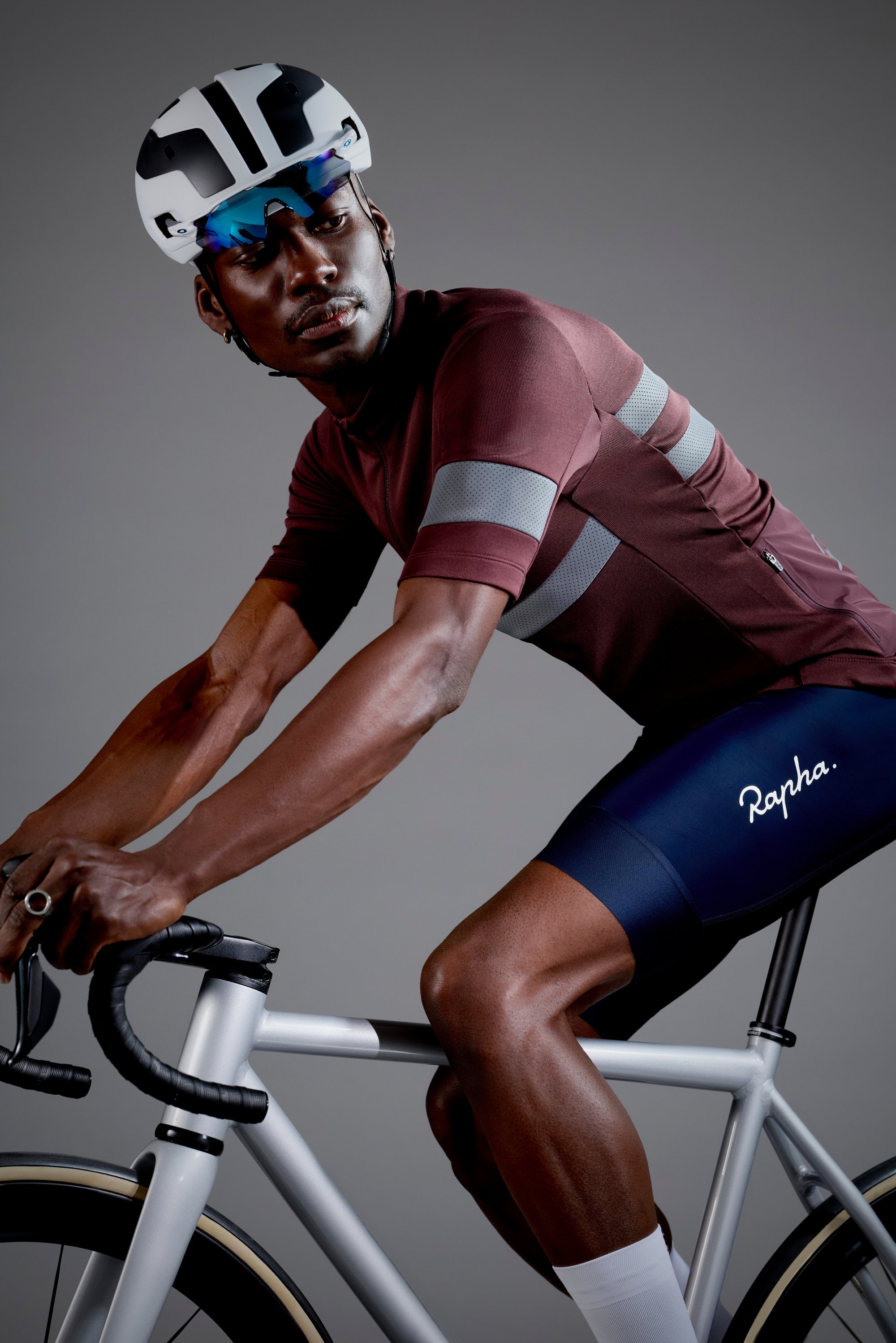 Cyklisti i maroon triko og svartshorts lener seg fremover på en elegant sølvveissykkel, iført hvit hjelm og solbriller.