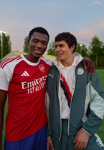 Deux jeunes hommes souriants sur un terrain de sport, l'un portant un maillot d'Arsenal, l'autre une veste de Manchester City, s'enlaçant.