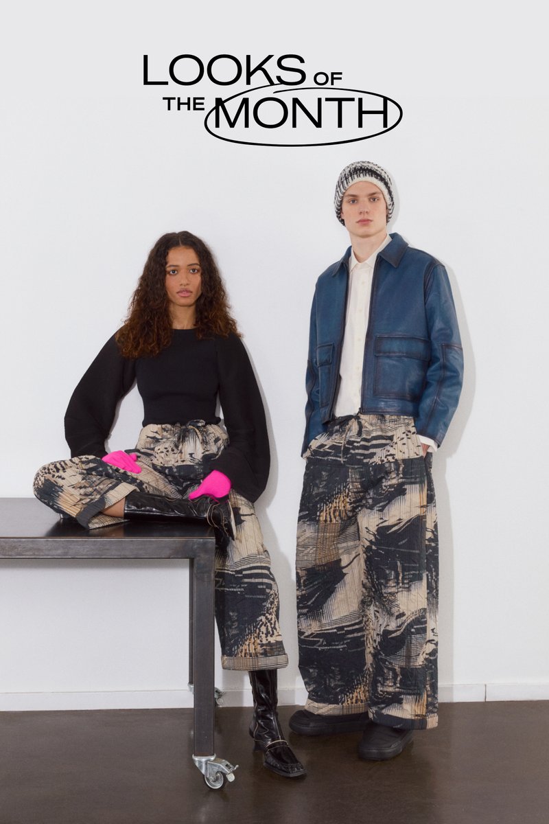 Dos modelos están de pie contra una pared sencilla. Uno está sentado en una mesa de metal, vistiendo una blusa negra y pantalones estampados, mientras que el otro lleva una chaqueta de cuero.