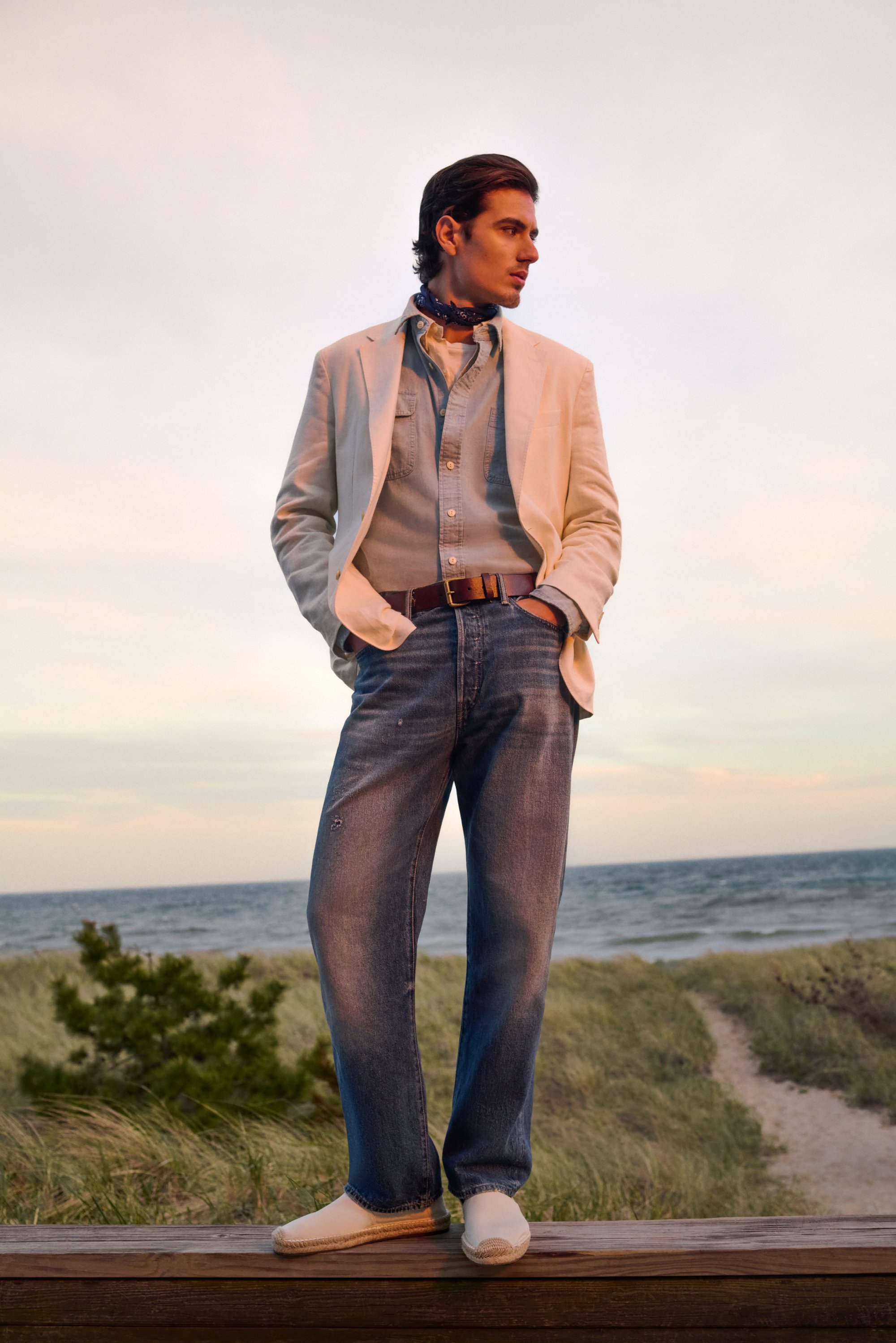 Un hombre está de pie en una plataforma de madera, vistiendo un blazer claro, una camisa de denim y jeans, con un fondo de playa y océano al atardecer.