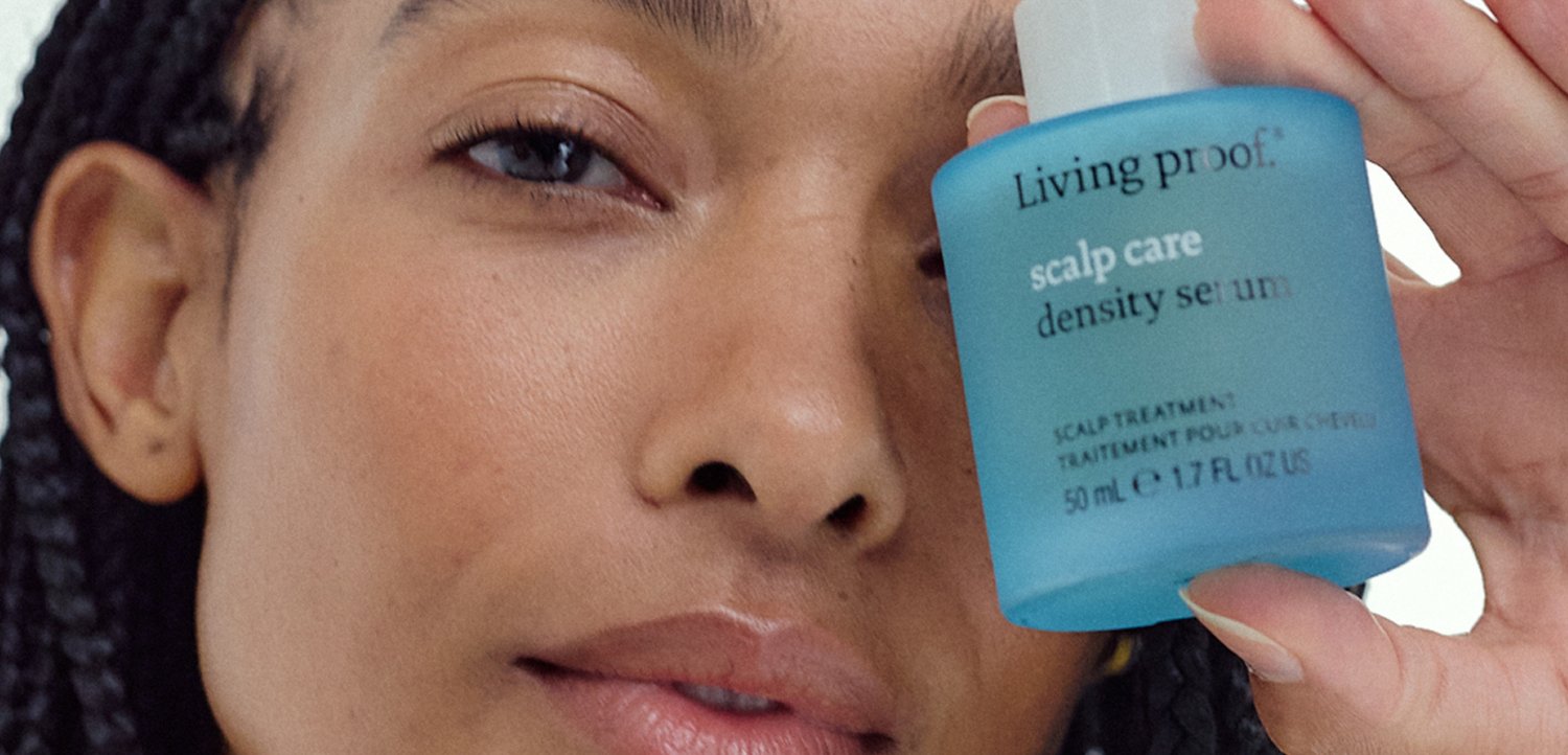 Frau hält eine blaue Flasche des Living Proof Kopfhautpflege-Dichte-Serums nah an ihr Gesicht und konzentriert sich auf das Produktetikett.