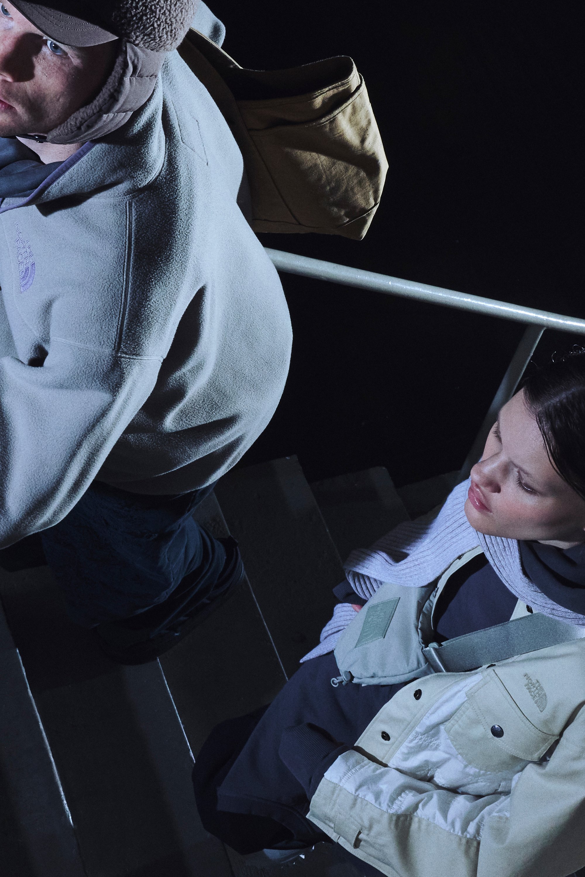 Due persone salgono le scale di notte. Un uomo con un cappello marrone e una giacca di pile porta una borsa. Una donna indossa abiti a strati e appare contemplativa.