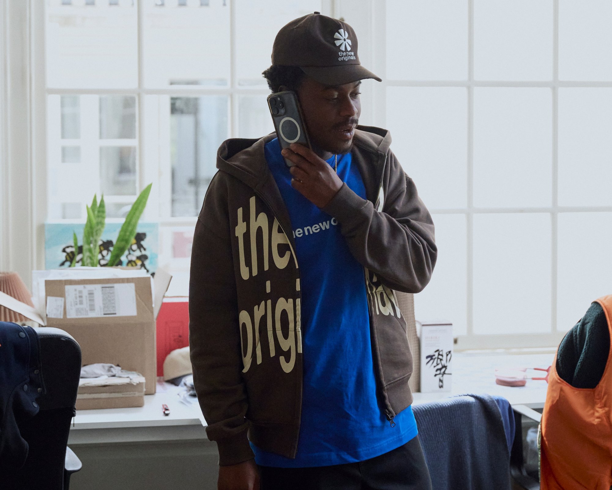 Hoodie zippé marron avec le texte "l'original", t-shirt bleu, tenant un smartphone noir, à l'intérieur avec des plantes et des boîtes en arrière-plan.