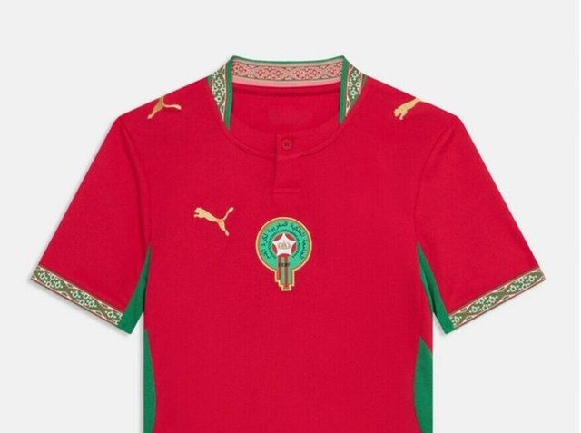 Piros, rövid ujjú Puma sportmez, zöld és arany mintás gallérral és mandzsettával, zöld kör alakú jelvénnyel, amelyen arab írás és korona látható.