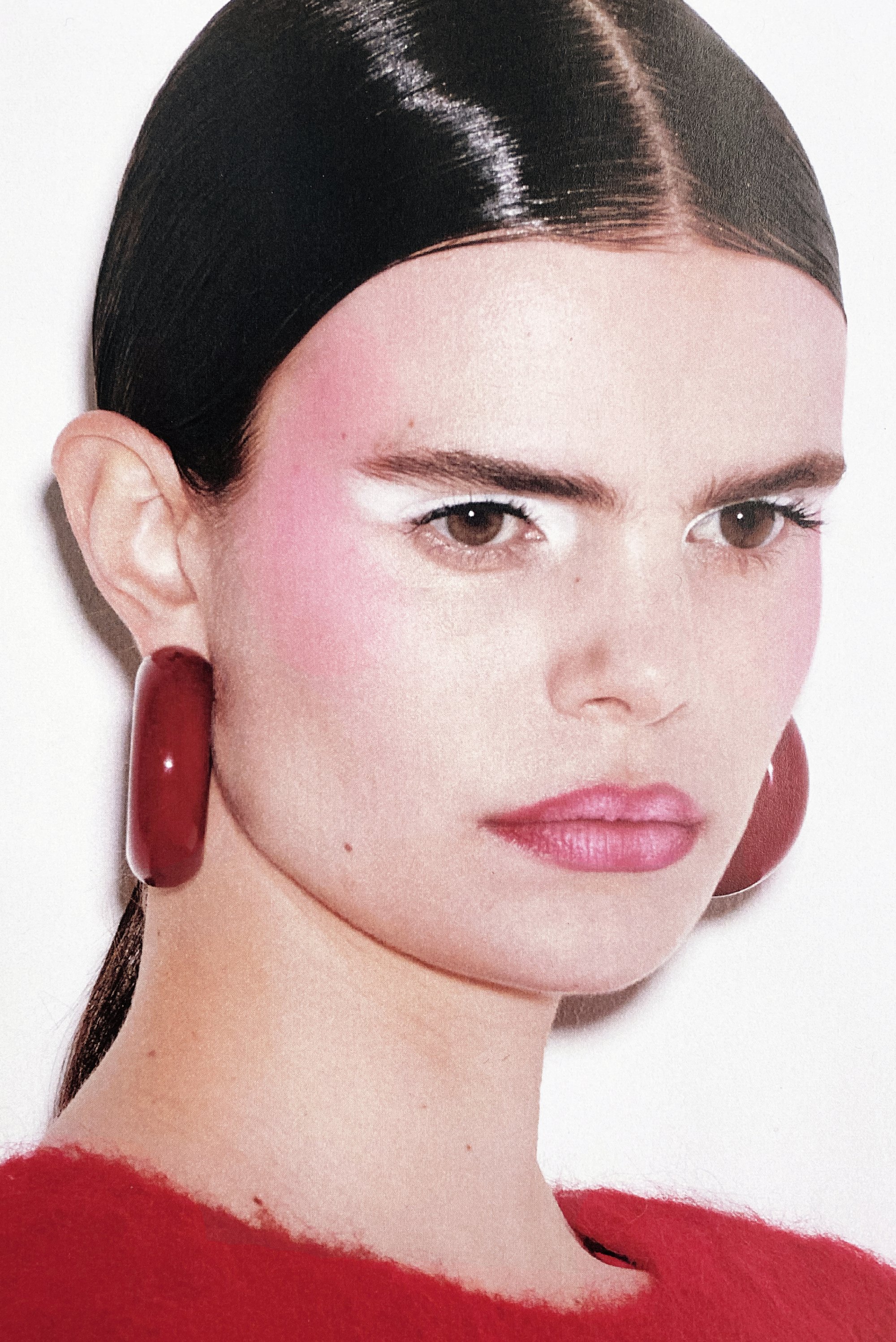 Modella con capelli neri lucidi, blush rosa audace e ombretto bianco, che indossa grandi orecchini a cerchio rossi e un top morbido rosso. Espressione chiara.