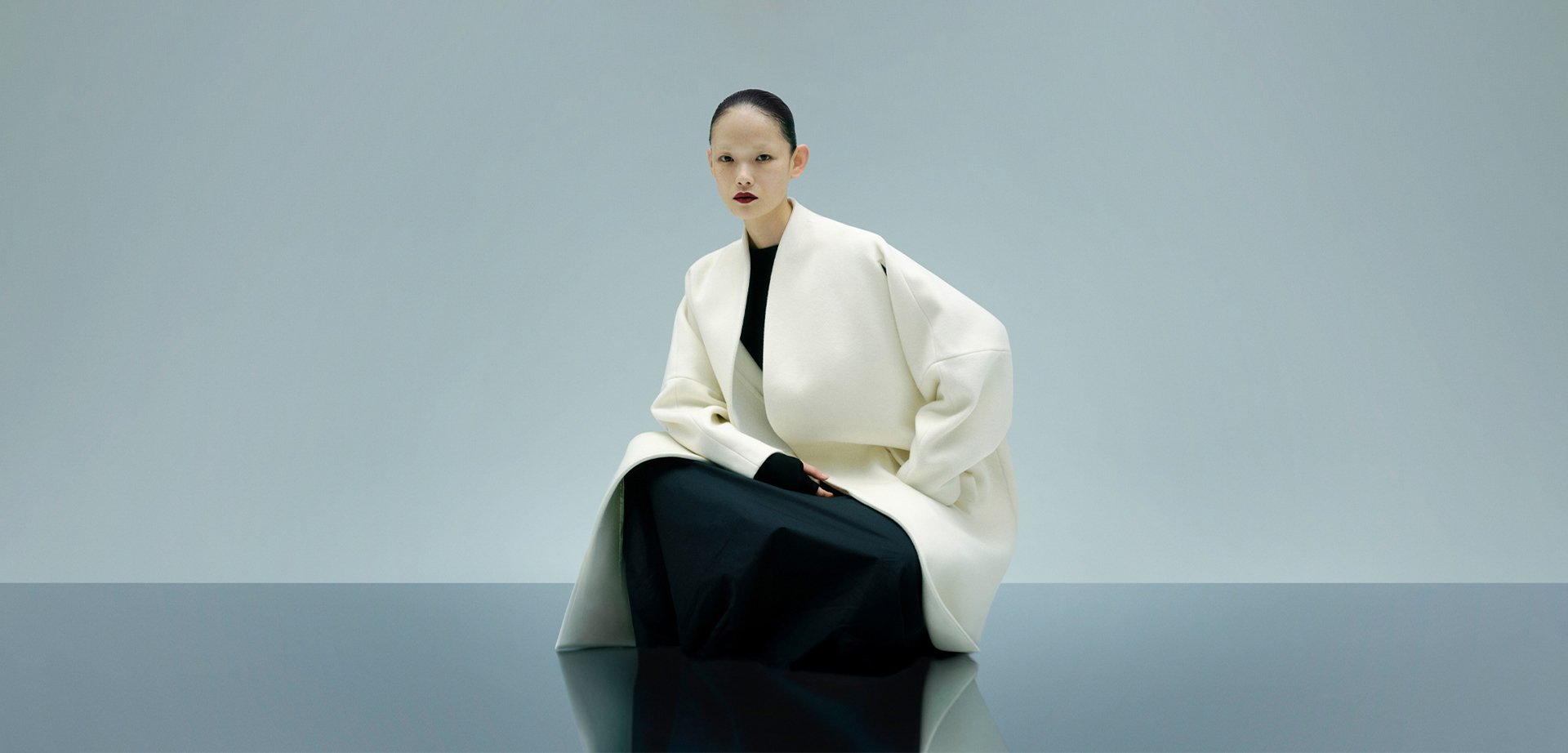 White oversized coat with a structured design, worn over a black garment. Smooth texture, minimalist style, and a reflective surface underneath.