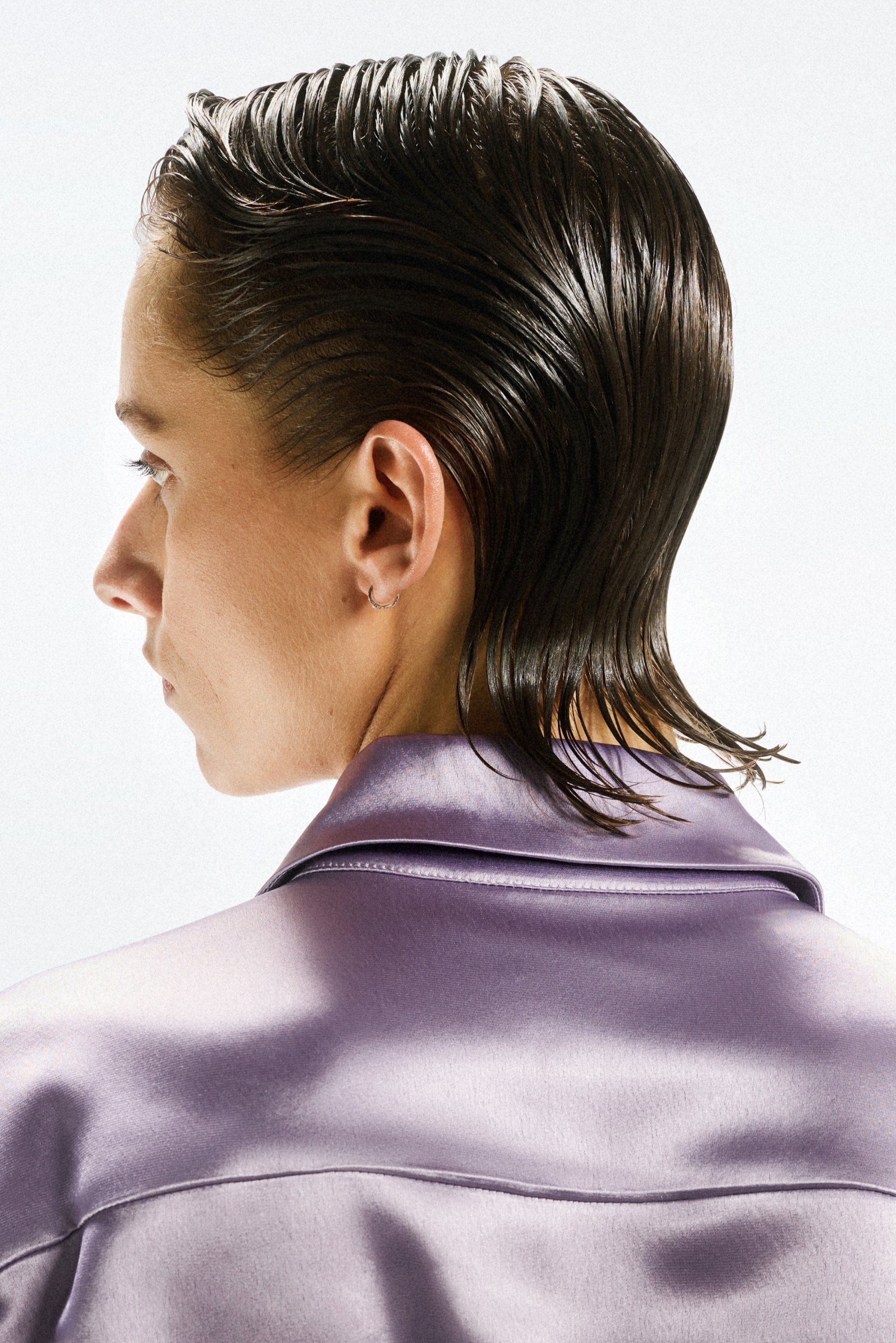 Een persoon met naar achter gekamd haar draagt een paarse satijnen blouse, gezien van achteren, waarbij hun profiel en haarstijl worden benadrukt.