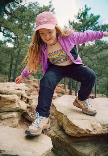 Junge Person in Outdoor-Bekleidung und Wanderschuhen, die vorsichtig über große Felsformationen in einem bewaldeten Gebiet geht.